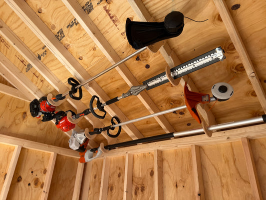 Three cordless string trimmers and a leaf blower hanging from a metal ceiling rack inside a wooden garage or shed with exposed wood framing.